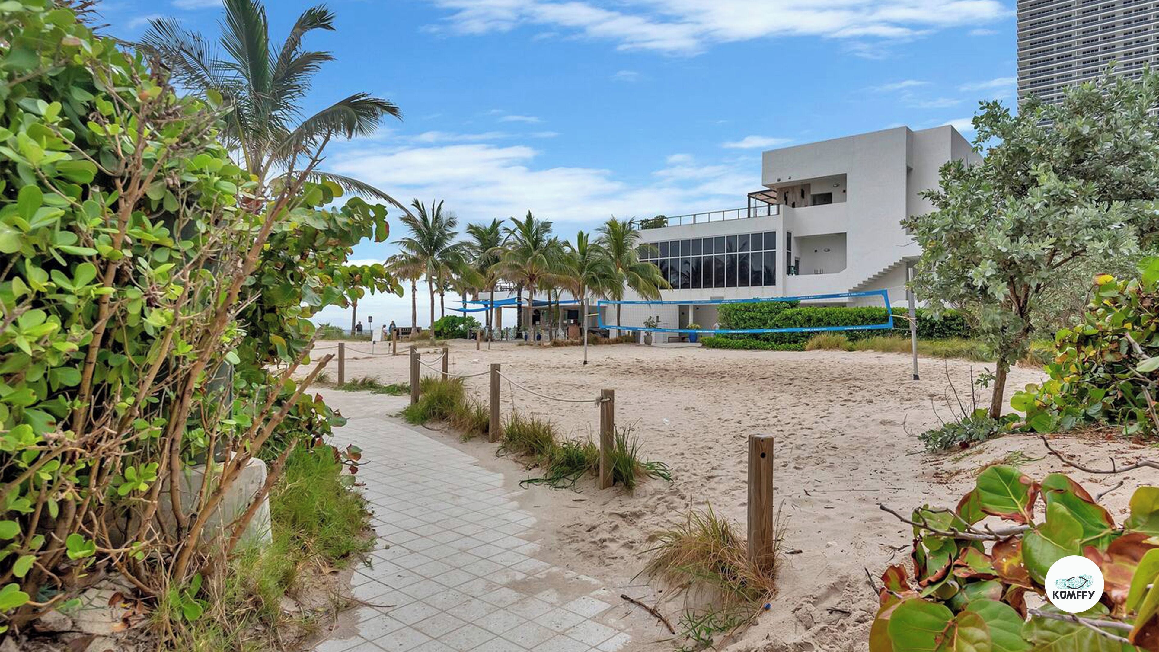 A pathway leading to a modern building with a beach view, surrounded by palm trees and greenery.