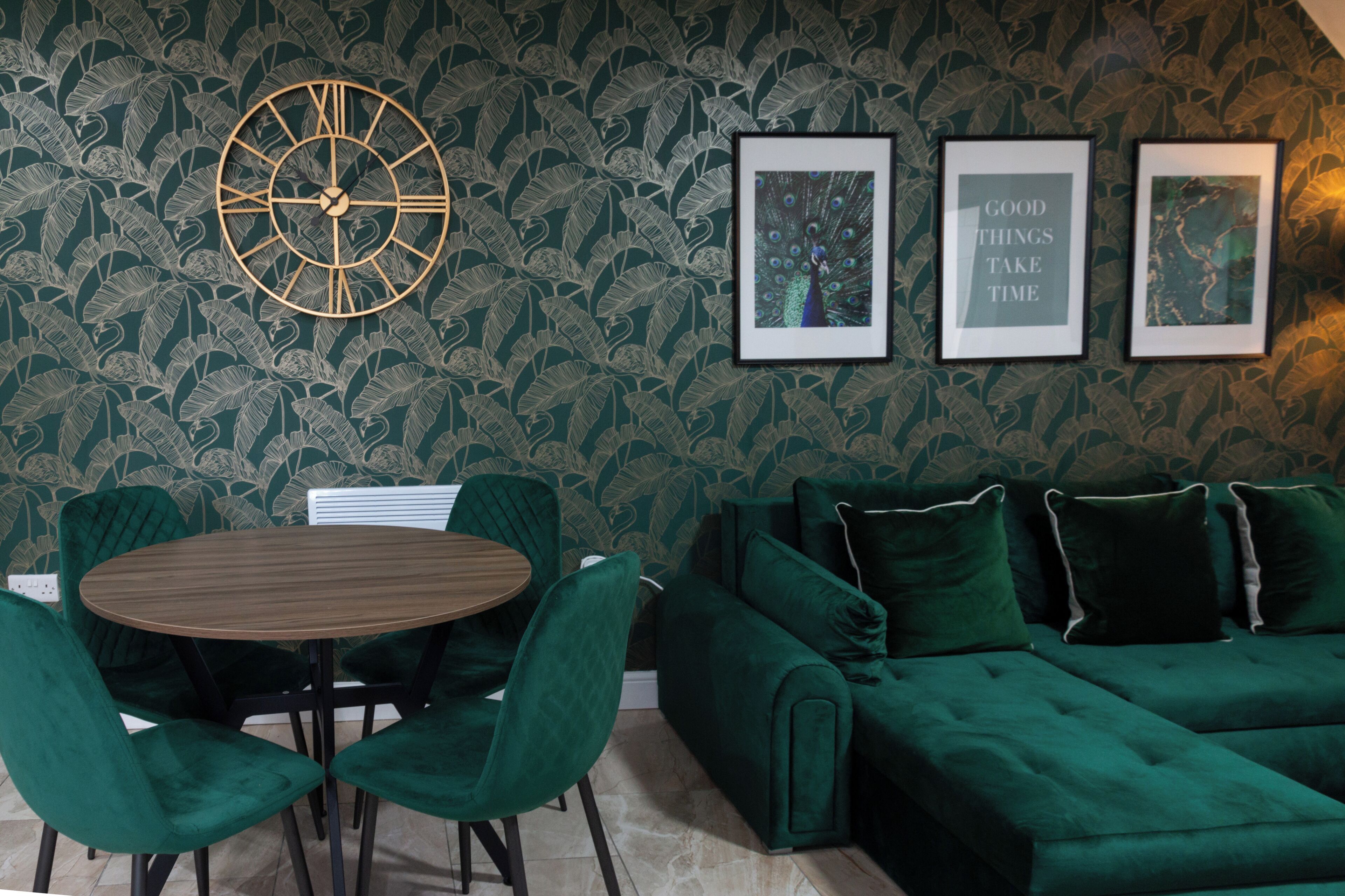 Une pièce avec un papier peint à motifs verts, une table ronde en bois entourée de chaises, un canapé vert et des tableaux encadrés accrochés au mur.