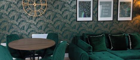 Une pièce avec un papier peint à motifs verts, une table ronde en bois entourée de chaises, un canapé vert et des tableaux encadrés accrochés au mur.