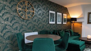 A modern living room with a green sofa, matching chairs, a round wooden table, and a large wall clock. - Velvet Luxe Retreat Opposite Stadium (Cardiff)