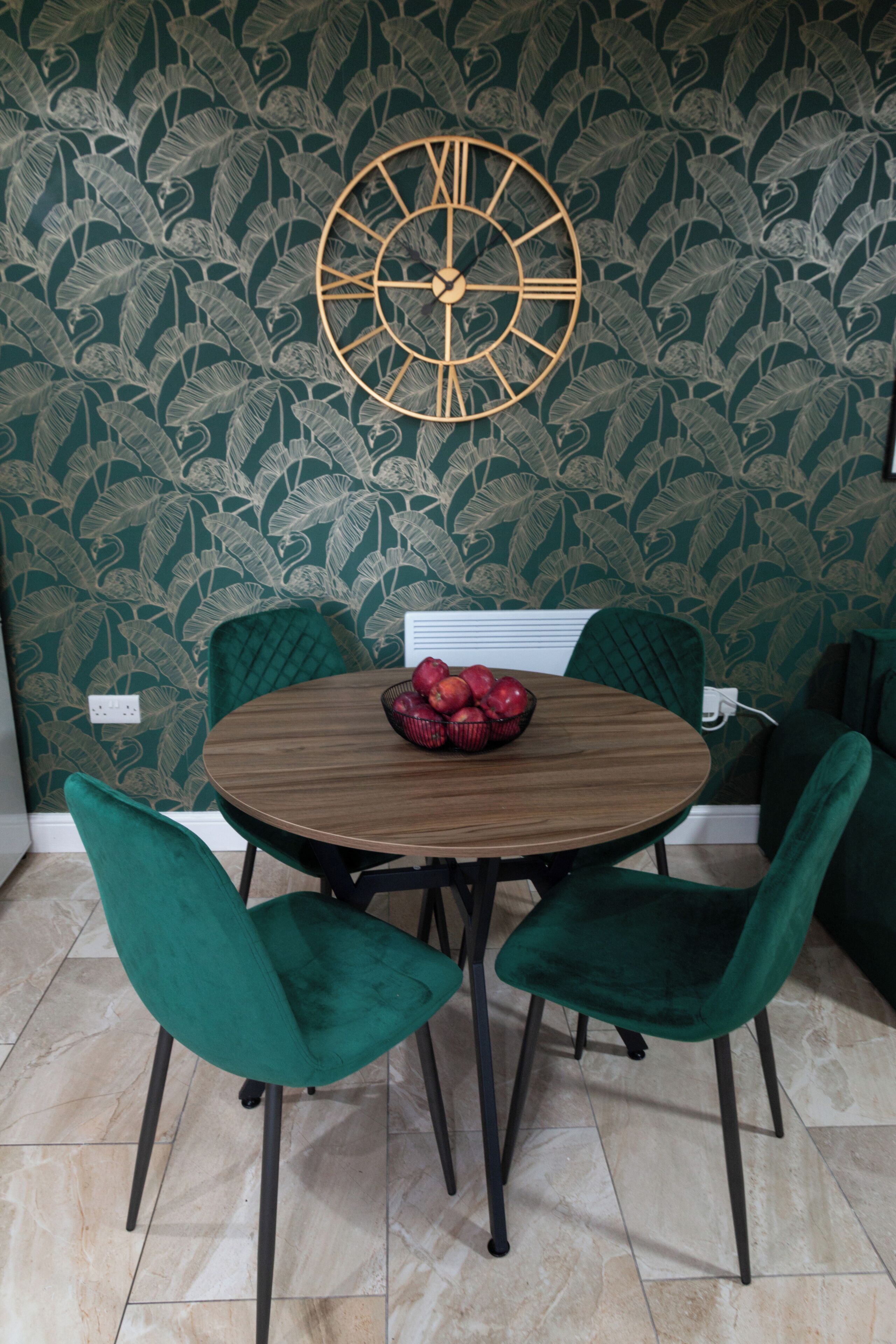 Un coin repas avec une table ronde en bois, deux chaises en velours vert et une horloge murale.