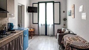 A compact kitchen with a stove, sink, and blue cabinet. A dining area with a patterned table and chairs is visible. The room has a large window with curtains and a view of the outside. - House at the Marina 1 (Trapani)