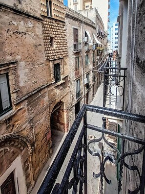 Une rue étroite en milieu urbain, bordée de vieux bâtiments, de balcons et d’une rambarde en métal.