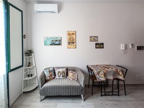 A room with a grey sofa, a small table with a patterned tablecloth, and wall decorations including a vintage car and a sign that says "Cheers".
