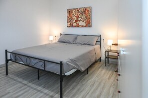 A single bed with a metal frame, grey bedding, two bedside tables with lamps, and a wall-mounted painting.