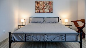 Un lit avec un cadre en métal, deux tables de chevet avec des lampes, une chaise en bois et un tableau abstrait aux couleurs vives accroché au mur.