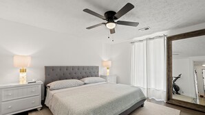 Une chambre à coucher avec une tête de lit capitonnée, un lit recouvert d’une couvre-lit à motifs, des lampes de chevet, un ventilateur de plafond et un grand miroir.