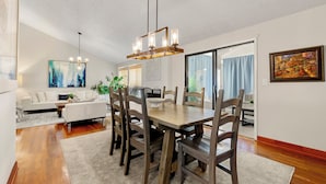 Un coin repas avec une table et des chaises en bois, un lustre et une vue sur un salon comprenant un canapé blanc et un tableau.