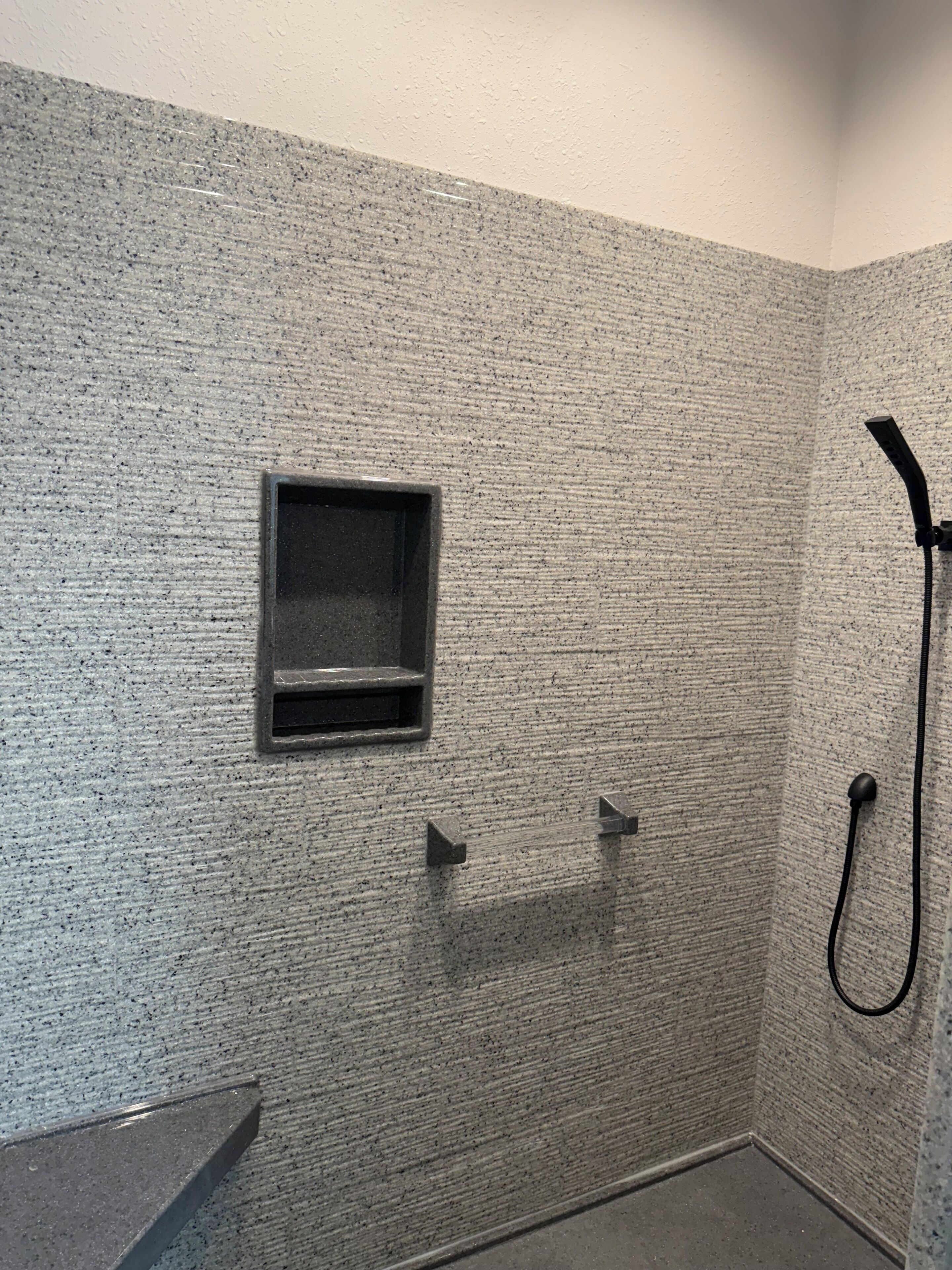 A small, enclosed shower area with a textured wall, a built-in shelf, and a handheld showerhead.