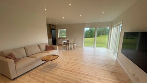 A modern living room with a sofa, coffee table, and a TV. There is a dining area with chairs and a table. The room has wooden flooring and large windows. - Farm idyll with lake views, private beach spot & boat near the west coast (Västra Götalands län)