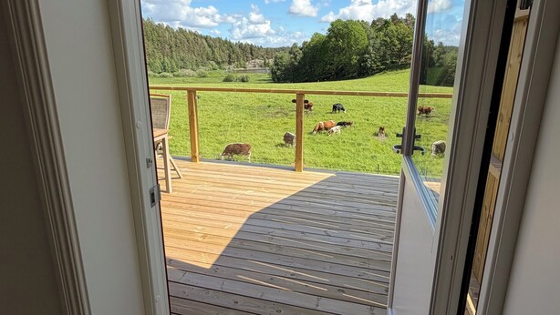 An open door leading to a wooden deck with a view of a grassy field and trees. - Farm idyll with lake views, private beach spot & boat near the west coast (Västra Götalands län)