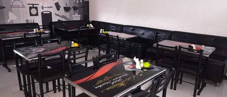 A restaurant interior with multiple tables and black chairs, a wall with kitchen utensils and a sign, and a clean tiled floor.