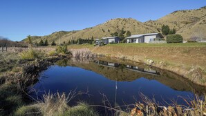 A serene landscape with a pond reflecting a modern house and a hill in the background. - Hurunui River Retreat - Tussock Cottage (Canterbury)