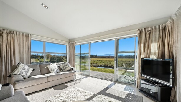 A modern living room with a grey sofa, a flat-screen TV, and large windows offering a view of a landscape. - Hurunui River Retreat - Tussock Cottage (Canterbury)