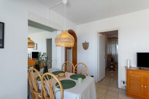 Un coin repas avec une table dressée pour quatre personnes, entourée de chaises en bois, d’un luminaire suspendu et d’un téléviseur posé sur un meuble en bois.
