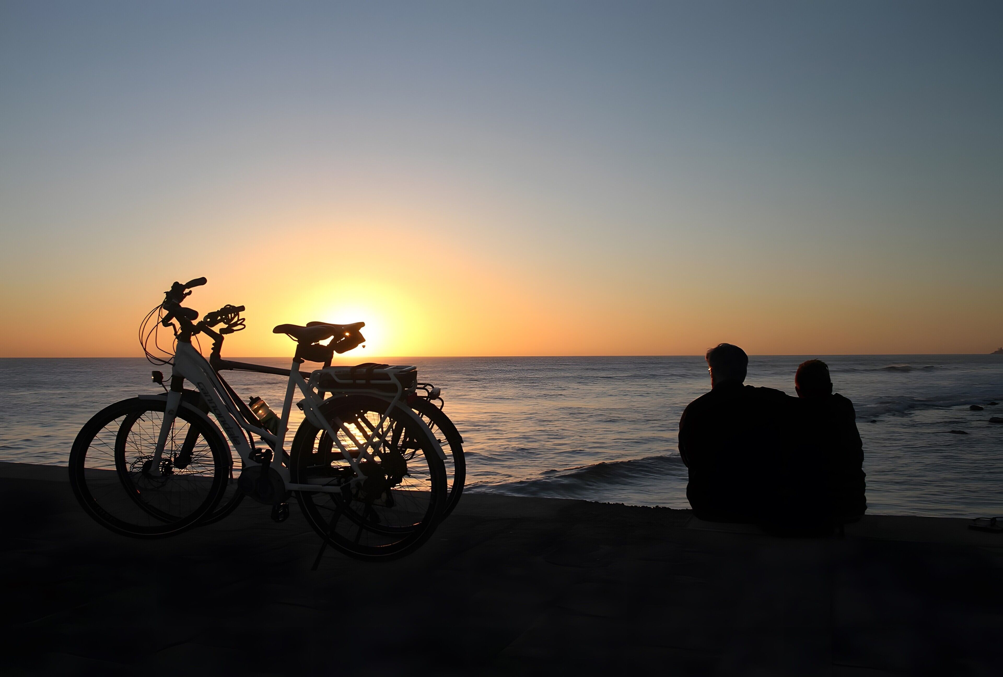 A sunset over the ocean with a bicycle and two people sitting on the beach.