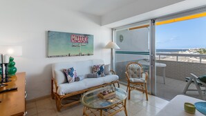 Ein Balkon mit Blick auf den Strand, eine weiße Couch mit Kissen, ein Korbstuhl, ein Glastisch und ein Gemälde eines Motels.