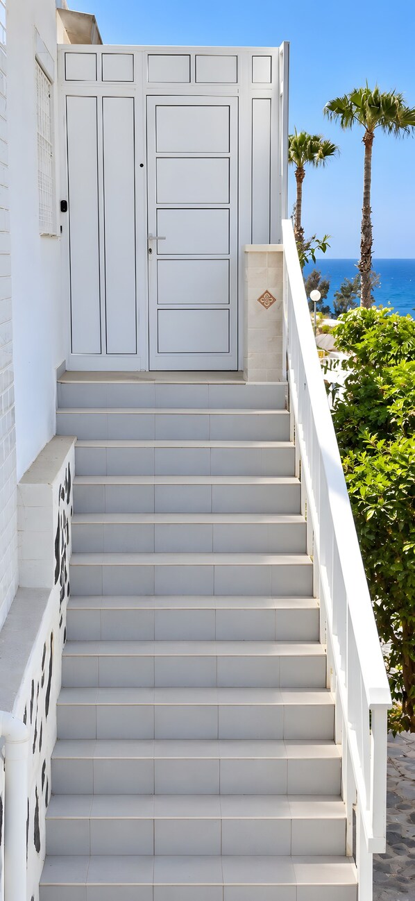 A set of white stairs leading to a door with a keypad lock, surrounded by a white railing, with a view of the sea and palm trees in the background.