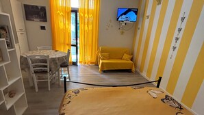 A room with a bed featuring a butterfly print, a yellow sofa, a TV mounted on the wall, and a dining area with a table and chairs.