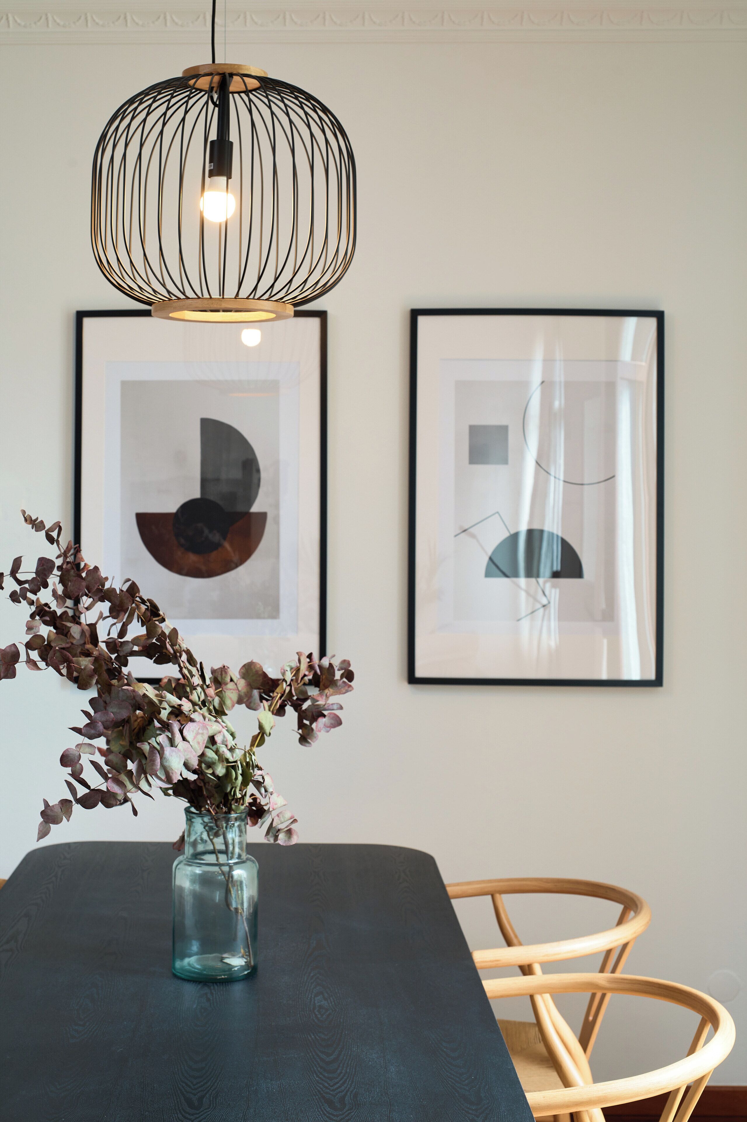 A dining area with a dark table, a vase with dried flowers, a hanging light fixture, and two framed abstract artworks on the wall.