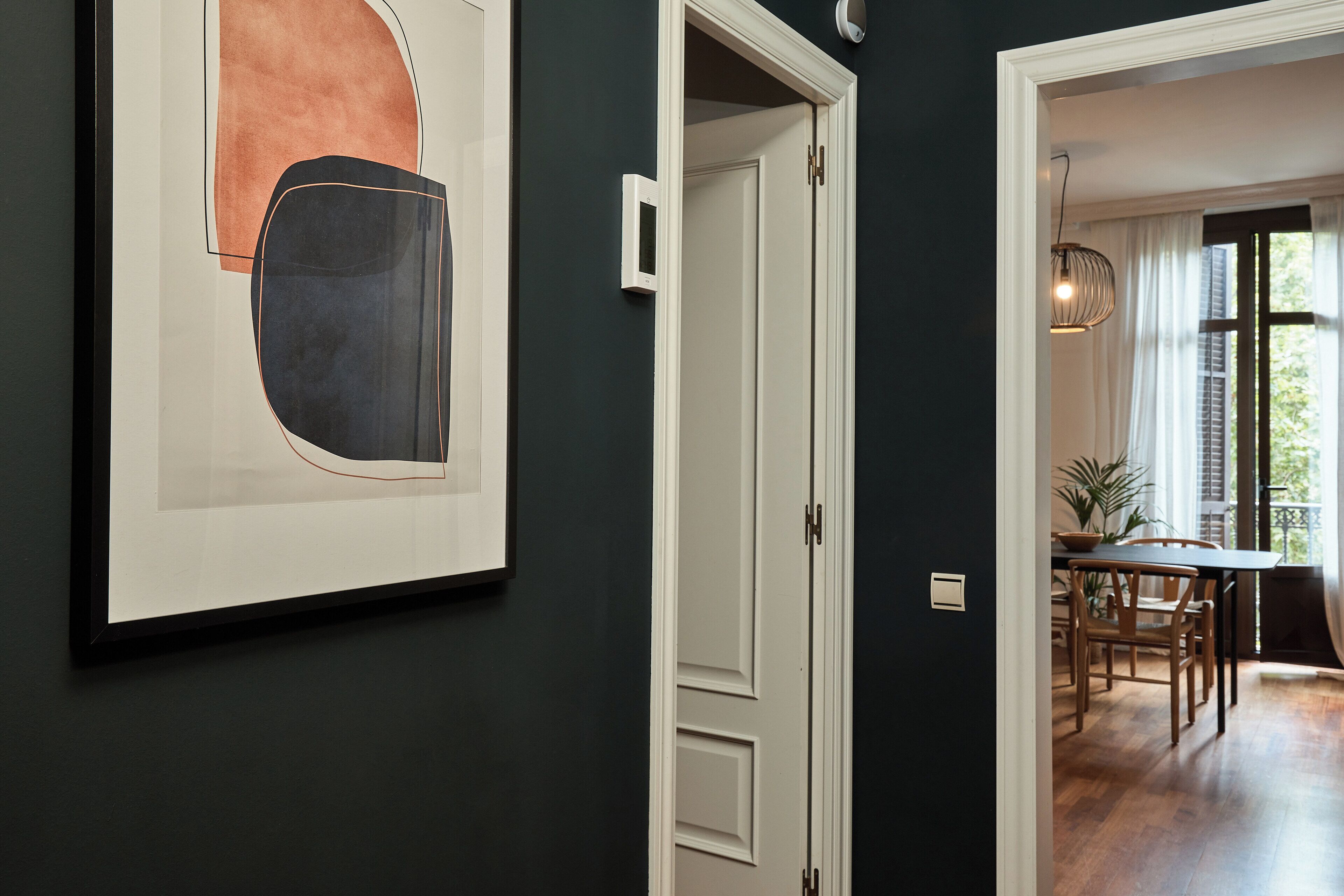 Un couloir orné d’un tableau abstrait encadré, une porte blanche et un coin repas avec des meubles en bois et un luminaire suspendu.