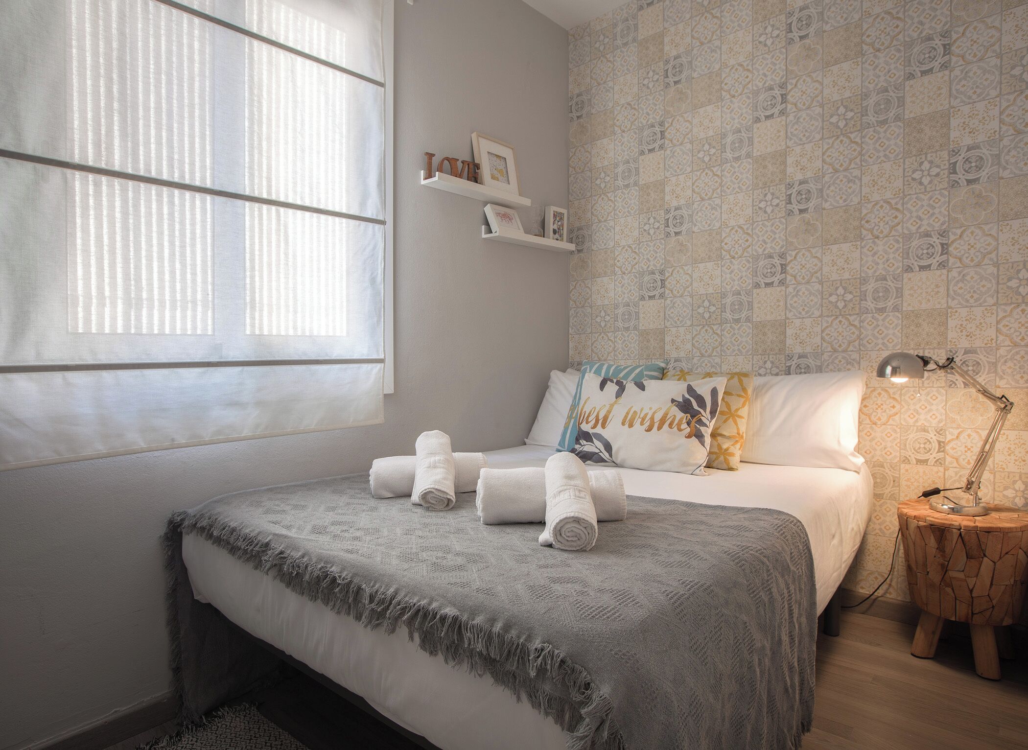Un dormitorio con cama, una ventana con persianas, una mesita de noche con lámpara y una pared con azulejos decorados.