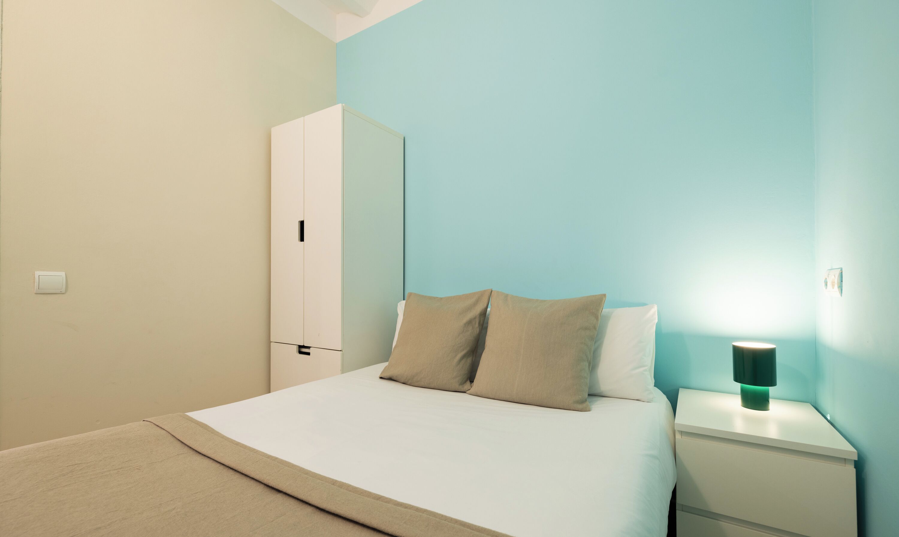 A bedroom with a bed, two pillows, a nightstand with a lamp, and a wardrobe.
