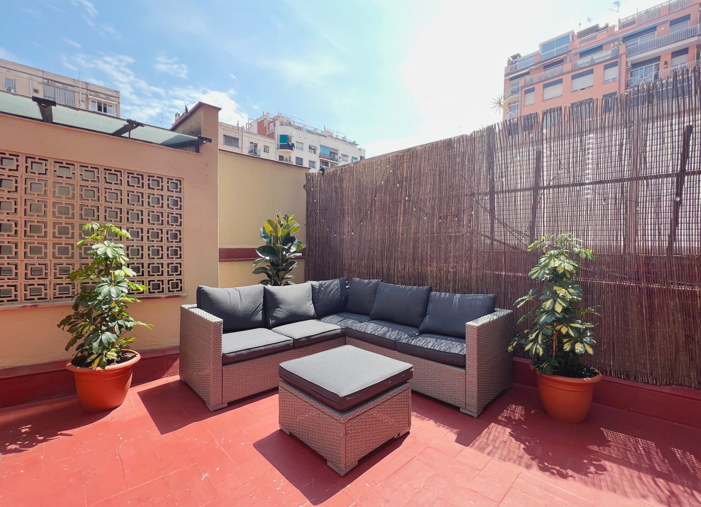 Une terrasse sur le toit, aménagée avec un canapé modulable et un pouf, entourée de plantes en pot et d’une clôture en bambou.
