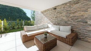A modern outdoor living area with a wicker sofa and a glass coffee table, set against a stone wall and a view of a green garden. - Stay U-nique Apartment Romana (Marbella)