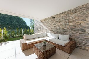 Een moderne buitenruimte met een rieten bank en een glazen bijzettafeltje, gelegen tegenover een stenen muur en een uitzicht op een groene tuin.