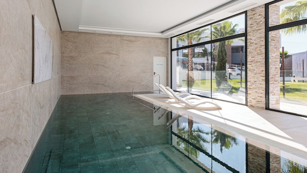 A modern indoor swimming pool with clear glass walls, a lounge area with white chairs, and a view of palm trees and buildings outside. - Stay U-nique Apartment Romana (Marbella)