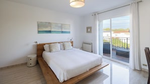Une chambre avec un grand lit, une œuvre d’art représentant un paysage urbain, un radiateur et une vue sur un balcon avec une balustrade.