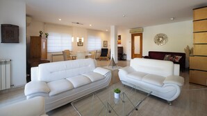 Un salon moderne avec deux canapés blancs, un coin repas avec une table et des chaises, et un espace cuisine équipé d’une cuisinière et de placards.