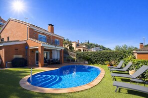 Una piscina rodeada de un césped cuidado, un patio con mobiliario exterior y una casa de ladrillo con balcón.