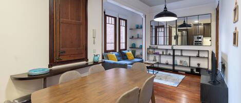 A dining area with a wooden table and chairs, a sofa with cushions, and a kitchen area with shelves and a refrigerator.