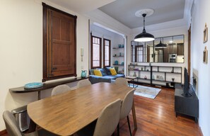 Un comedor con mesa y sillas de madera, un sofá con cojines y una zona de cocina con estantes y un refrigerador.