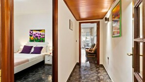 A hotel room with a bed, a nightstand, a painting, and a hallway leading to a sitting area.
