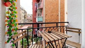 A balcony with a wooden table and benches, a hanging flower arrangement, and a view of adjacent buildings.