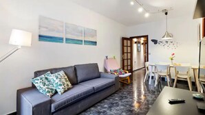A modern living room with a grey sofa, a dining table with chairs, and a small kitchen area.