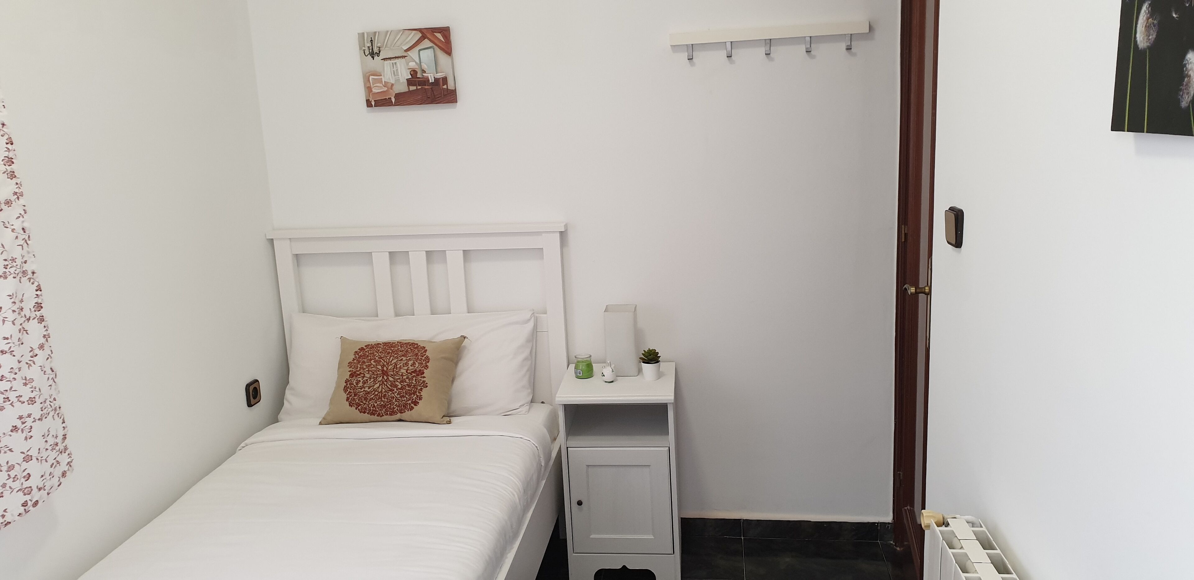 Un dormitorio pequeño y blanco con una cama individual, una mesita de noche, un estante pequeño y una puerta.