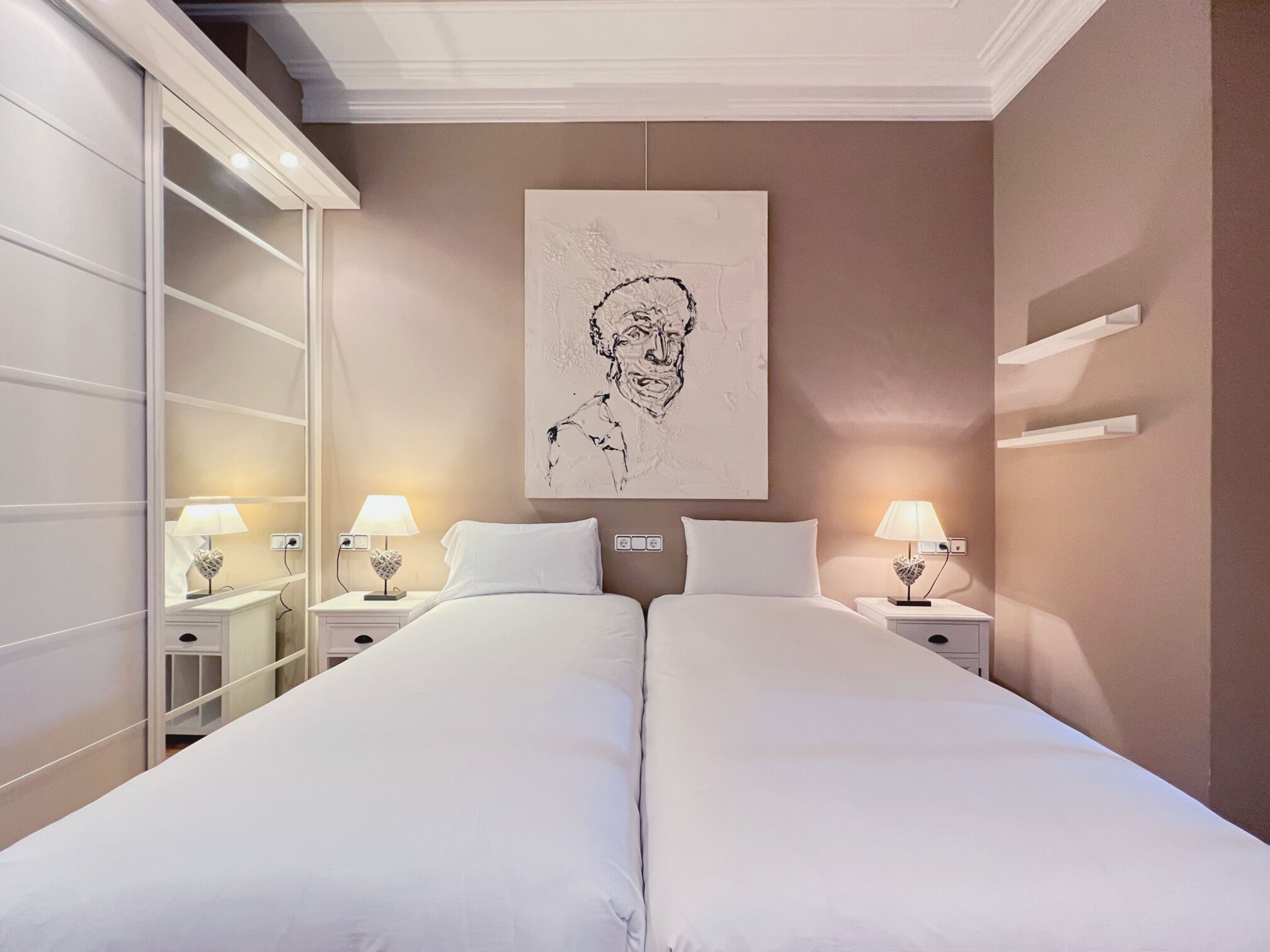A double bed with a white duvet, two bedside tables with lamps, a large framed artwork on the wall, and a built-in wardrobe with sliding doors.