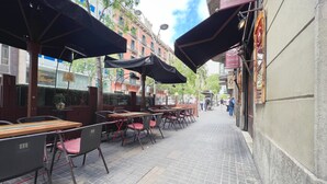 An outdoor café with tables and chairs, umbrellas, and a brick building in the background.