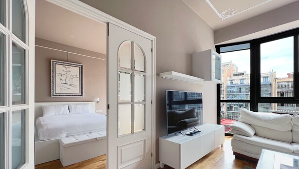 A modern hotel room with a bed, a TV, a sofa, and a large window offering a city view.