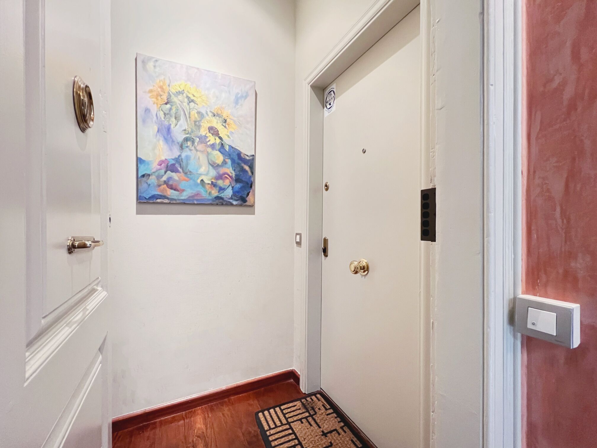 A hallway with a white door, a painting of flowers, and a wooden floor.