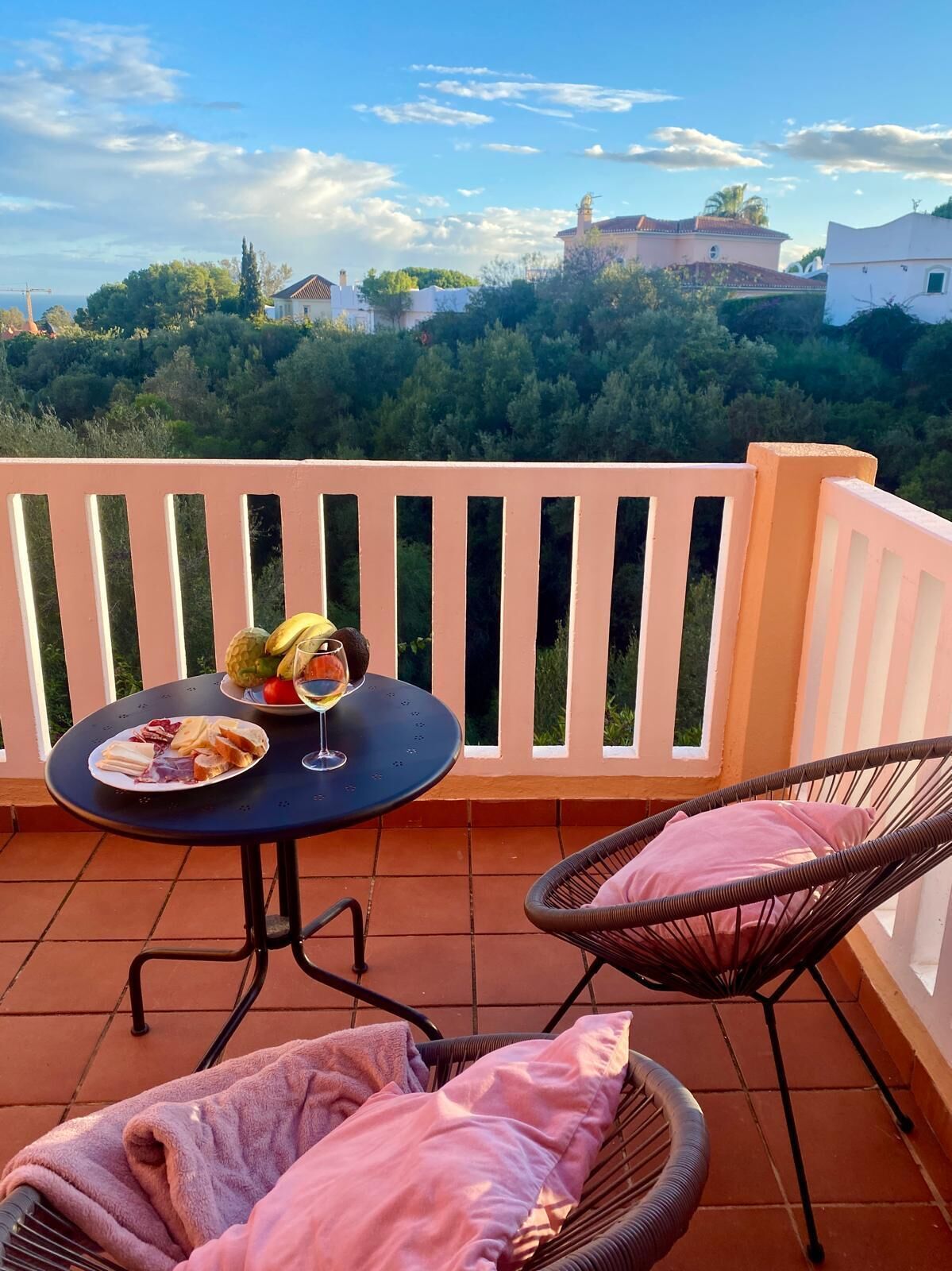 A balcony with a table set for a meal, two wicker chairs, and a view of a forested area with buildings in the distance.