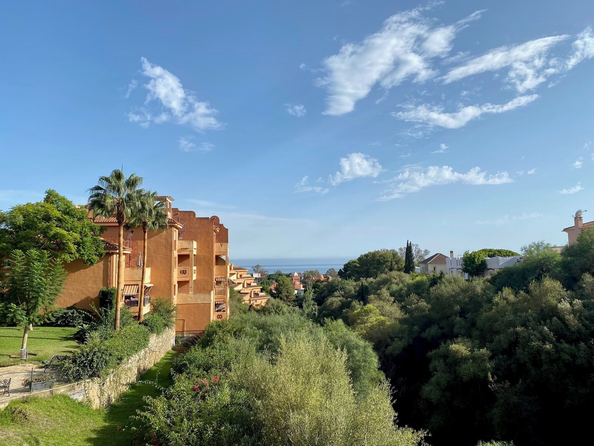 Zona residencial con un edificio de varios pisos que tiene balcones, rodeada de abundante vegetación y un cielo despejado.