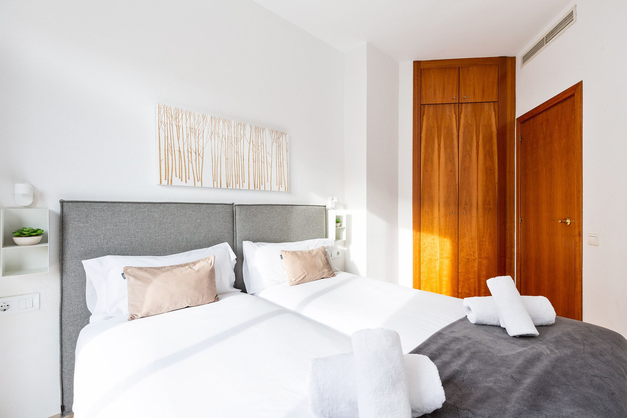 A neatly made bed with white and grey bedding, a grey headboard, and a wooden wardrobe beside it.