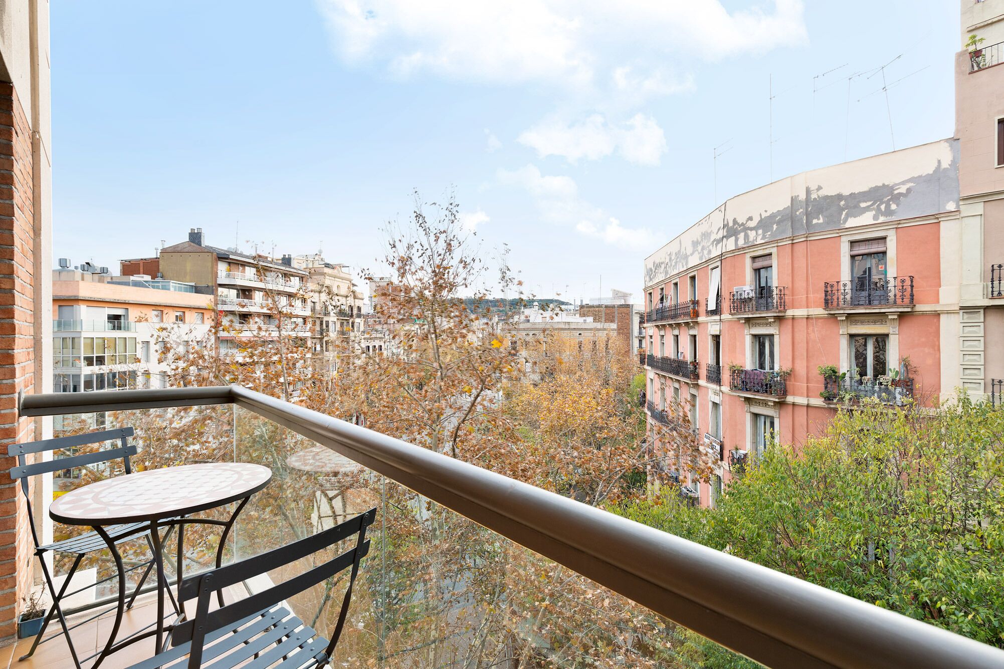 Un balcón con mesa y sillas, con vista a un paisaje urbano con edificios altos y árboles.