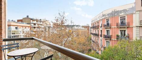 Un balcón con mesa y sillas, con vista a un paisaje urbano con edificios altos y árboles.