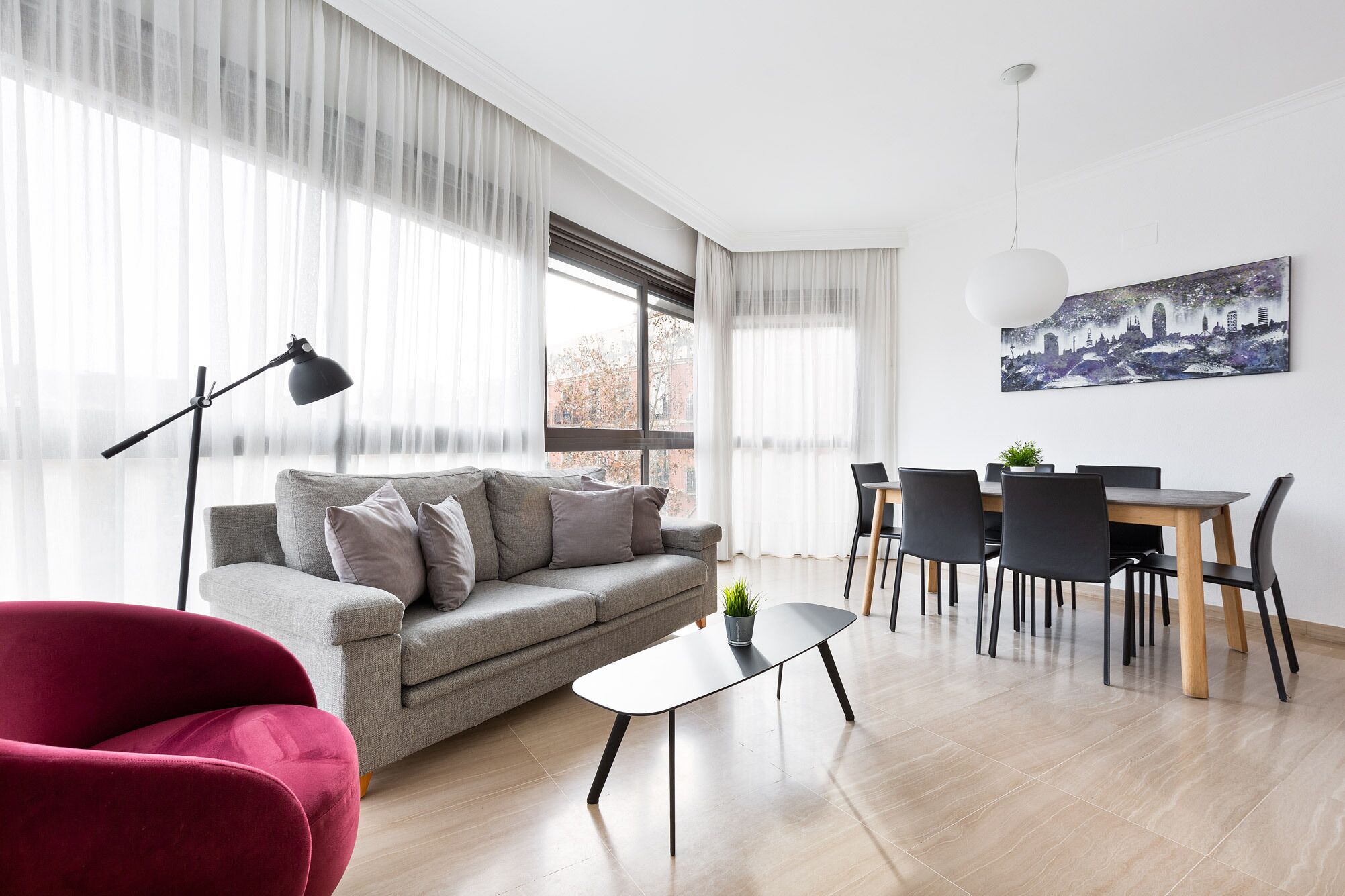 Una sala moderna con un sofá gris, un sillón rojo, una mesa de comedor con sillas y un cuadro de un paisaje urbano en la pared.